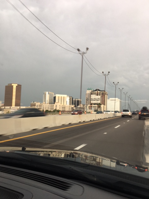 View of a city skyline from a highway, featuring tall buildings and traffic under a cloudy sky.