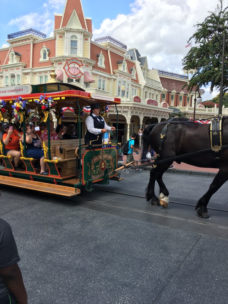 A vintage horse-drawn carriage with colorful decorations moves through a theme park, with dressed passengers inside, and a scenic backdrop of whimsical buildings and trees.