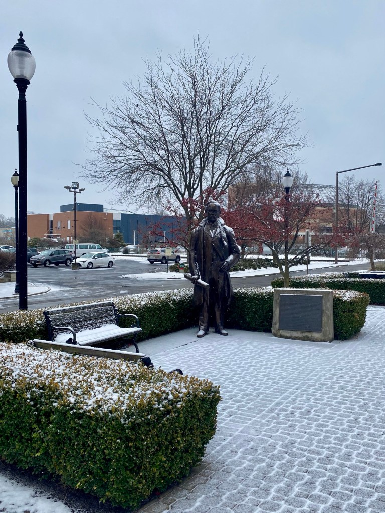 A bronze statue of a man in formal attire standing on a snow-covered sidewalk, surrounded by neatly trimmed hedges and a bench, with a lamppost and buildings in the background.