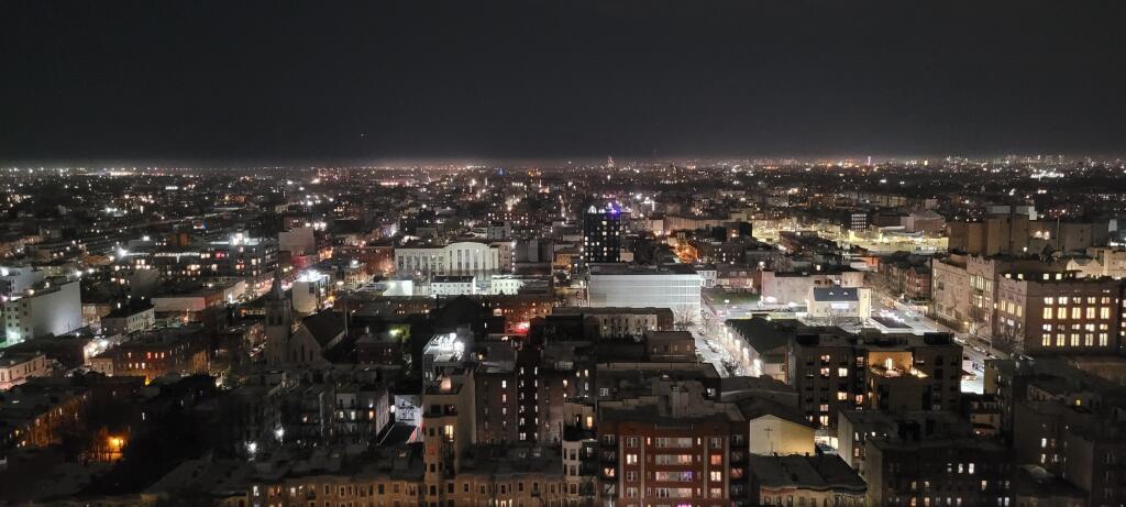A panoramic night view of a city skyline, showcasing numerous buildings illuminated by streetlights and neon signs, with a vast urban landscape extending into the horizon.